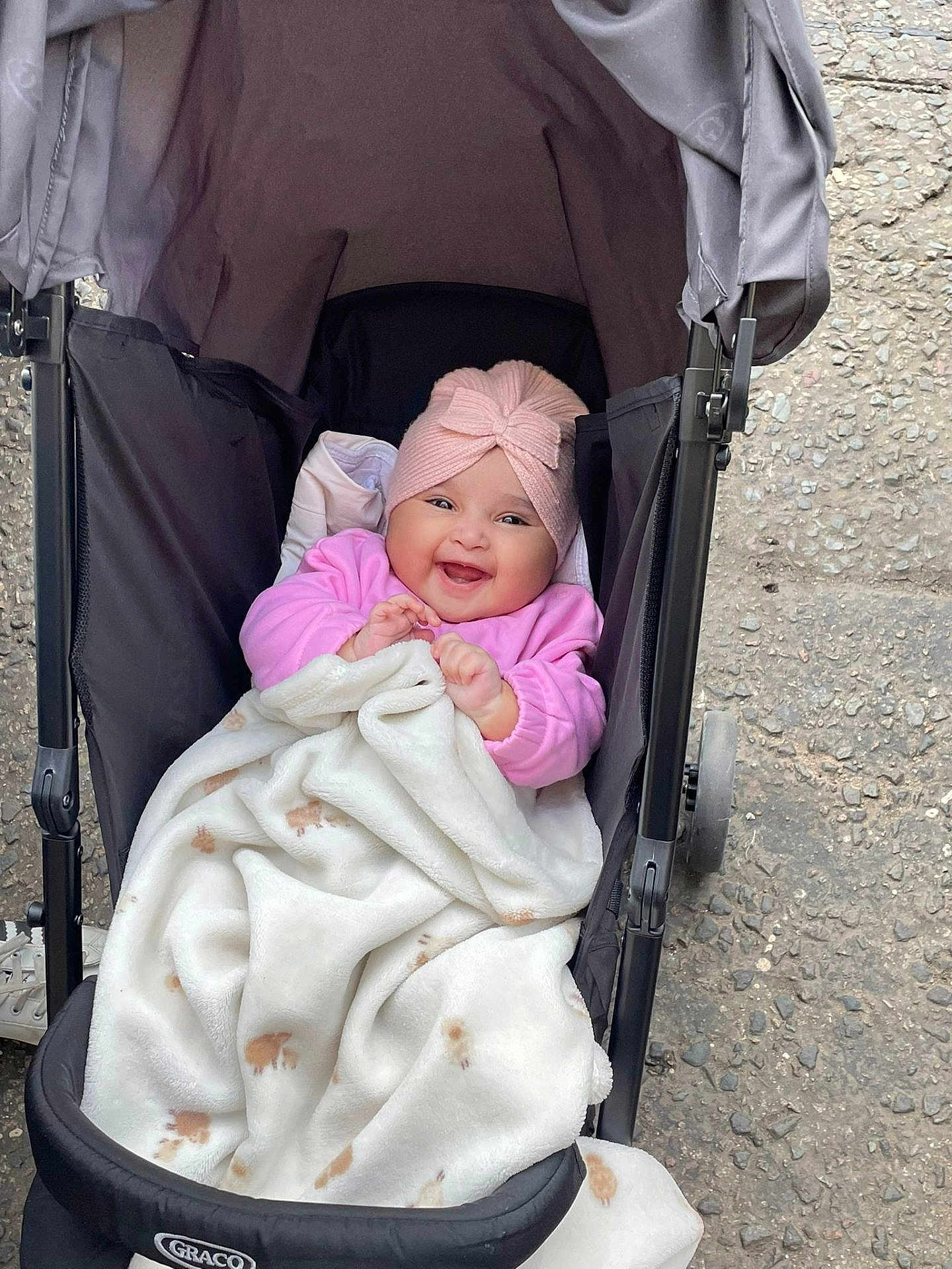 Ayzel is registered to the contest to win money with this photo: baby, stroller, blanket, pink_clothing, turban, smiling, happy, child, outdoor, pavement, black_stroller, cozy, wrapped, infant, cute, person, seat, graco_brand, warm, portrait