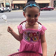 Isabella joined the competition — help win amazing prizes! child, girl, princess_dress, pink_dress, disney_princess, candy, headband, outdoor, sidewalk, table, red_table, city, buildings, cars, smiling, person, young_child, curly_hair, playful, cute