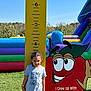 Isabella is registered to the contest to win money with this photo: child, boy, height_chart, applewood_orchards, cartoon_apple, inflatable, outdoor, grass, sunny, smiling, tshirt, short_sleeve, blue_pants, measurement, playground, fun, nature, trees, daylight, casual
