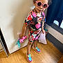 Isabella is registered to the contest to win money with this photo: child, girl, sunglasses, toy, pony, dress, colorful_clothing, wooden_floor, indoor, paper, wall, door, window, curtain, footwear, shoes, standing, person, smile, hair