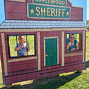 Isabella joined the competition — help win amazing prizes! child, adult, outdoor, sunlight, grass, playground, wooden_sign, mock_jail, window, bars, smile, person, fun, daylight, sunglasses, shirt, green_door, red_building, shadow, recreation