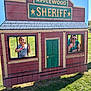child, adult, outdoor, sunlight, grass, playground, wooden_sign, mock_jail, window, bars, smile, person, fun, daylight, sunglasses, shirt, green_door, red_building, shadow, recreation