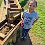 child, smiling, outdoor, grass, wood, water, play, sunlight, casual_clothing, tshirt, blue_pants, shoes, sand, wooden_structure, happy, person, daylight, nature, fun, playground