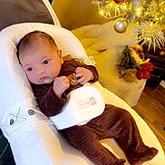 Jema participe au concours pour gagner de l'argent avec cette photo : baby, baby_seat, brown_pajamas, child, christmas_tree, cozy, cushion, cute, decorations, festive, furniture, holiday, holiday_lights, indoor, infant, ornaments, person, soft_lighting, warm_colors, wood_floor