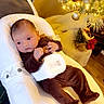 baby, baby_seat, brown_pajamas, child, christmas_tree, cozy, cushion, cute, decorations, festive, furniture, holiday, holiday_lights, indoor, infant, ornaments, person, soft_lighting, warm_colors, wood_floor