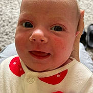Daisy is registered to the contest to win money with this photo: baby, infant, close_up, face, smile, eyes, hand, clothing, red_pattern, white_outfit, skin, head, person, young_child, carpet, indoors, cute, expression, happy, human