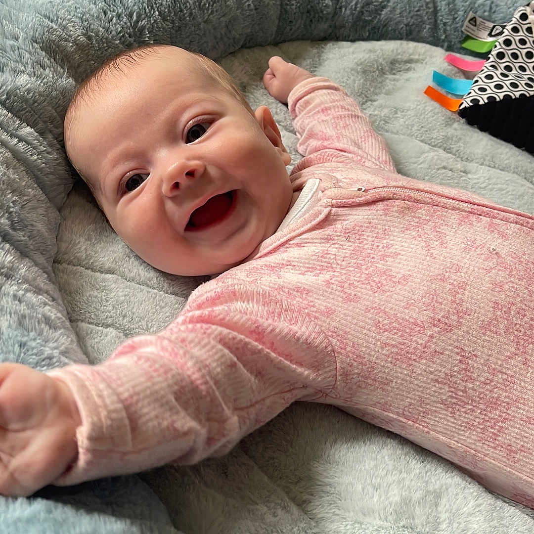 Daisy is registered to the contest to win money with this photo: arm_stretch, baby, baby_nest, bright_eyes, carpet, child, comfort, cozy, cute, face, happy, indoors, infant, lying_down, person, pink_clothing, playtime, plush, smiling, soft_toys