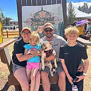 Pickles joined the competition — help win amazing prizes! family, dog, french_bulldog, child, adult, outdoor, bench, smiling, glasses, hat, tshirt, shorts, water_bottle, festival, tent, sunny, grass, wood, sign, sunglasses