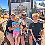 family, dog, french_bulldog, child, adult, outdoor, bench, smiling, glasses, hat, tshirt, shorts, water_bottle, festival, tent, sunny, grass, wood, sign, sunglasses