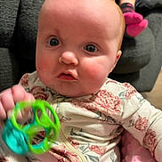 Adeline is registered to the contest to win money with this photo: baby, infant, baby_face, floral_onesie, toy, green_toy, rattle, closeup, portrait, indoor, sofa, pink_blanket, rosy_cheeks, big_eyes, red_hair, chubby_cheeks, hand, motion_blur, sitting, casual_clothing