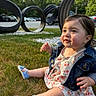 Mary is registered to the contest to win money with this photo: baby, child, grass, denim_jacket, blue_shoes, playground, outdoor, trees, parking_lot, daylight, curly_hair, cute, sitting, clothing, nature, park, toddler, face, smiling, shoes