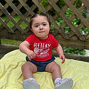 Mary is registered to the contest to win money with this photo: toddler, child, baby, sitting, outdoor, blanket, red_shirt, denim_shorts, pigtails, cute, wooden_lattice, greenery, curious, portrait, footwear, white_socks, casual_clothing, daylight, nature, expression