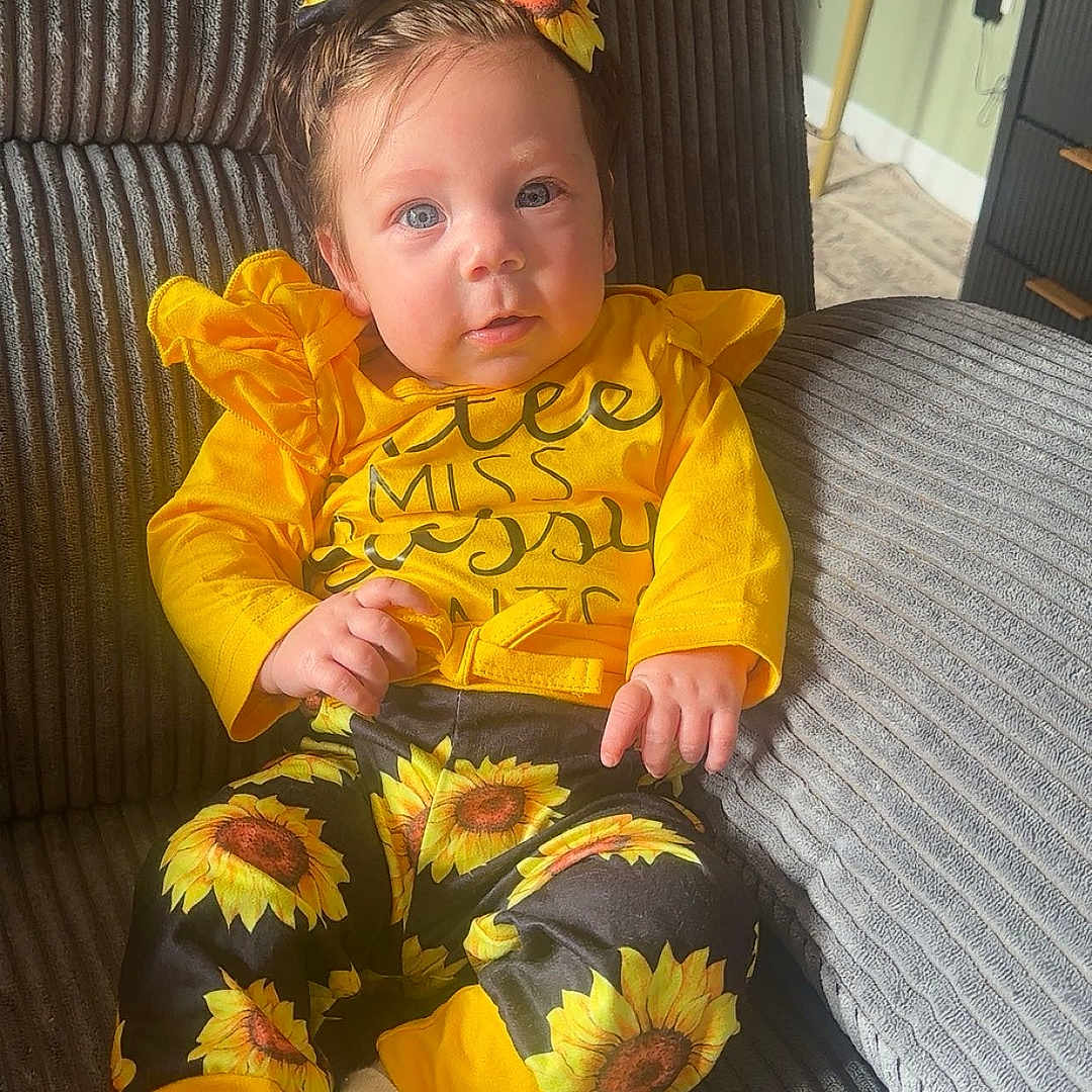 Everley is registered to the contest to win money with this photo: baby, bow, bright_colors, child, couch, cozy, cute, fashion, home, indoor, infant, patterned_pants, person, portrait, sitting, smiling, sunflower, white_socks, yellow_clothing, young_child