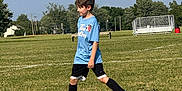 Odin joined the competition — help win amazing prizes! child, boy, soccer_ball, grass, soccer_field, blue_shirt, black_shorts, sports, outdoor, playing, cleats, trees, bench, bleachers, daytime, athlete, recreation, field, action, young
