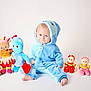 baby, child, blue_onesie, hood, plush_toys, stuffed_animals, sitting, smiling, indoor, white_background, cute, soft_toys, colorful, footwearless, happy, portrait, toddler, childrens_clothing, playtime, cozy