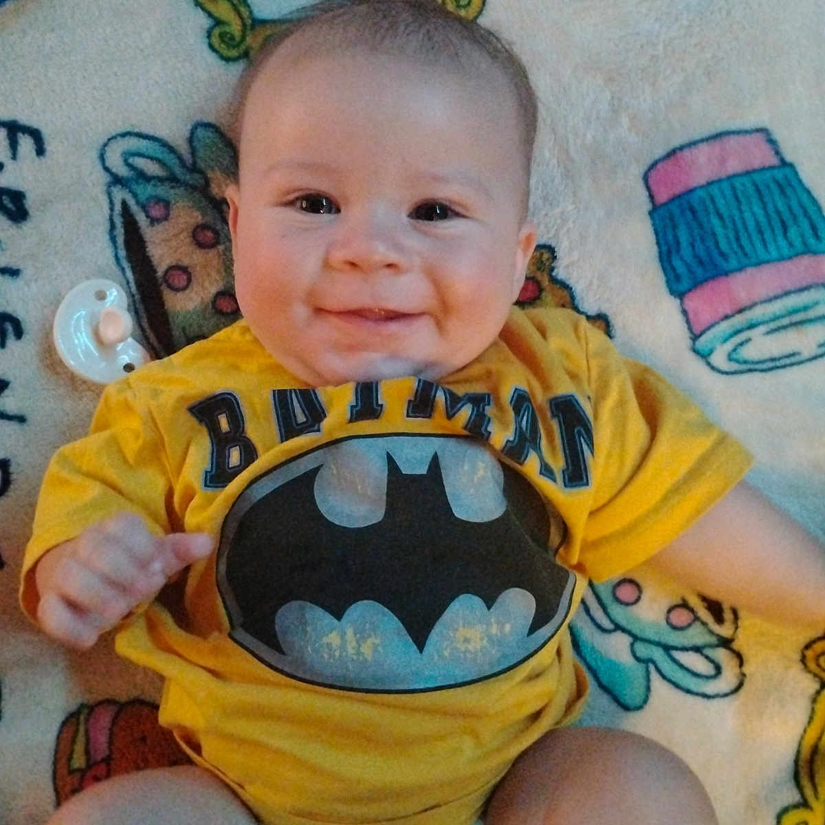 Riott is registered to the contest to win money with this photo: baby, batman, blanket, child, chubby_cheeks, clothing, cute, diaper, face, happy, head, indoor, infant, pacifier, person, playful, sitting, smile, yellow_shirt, young