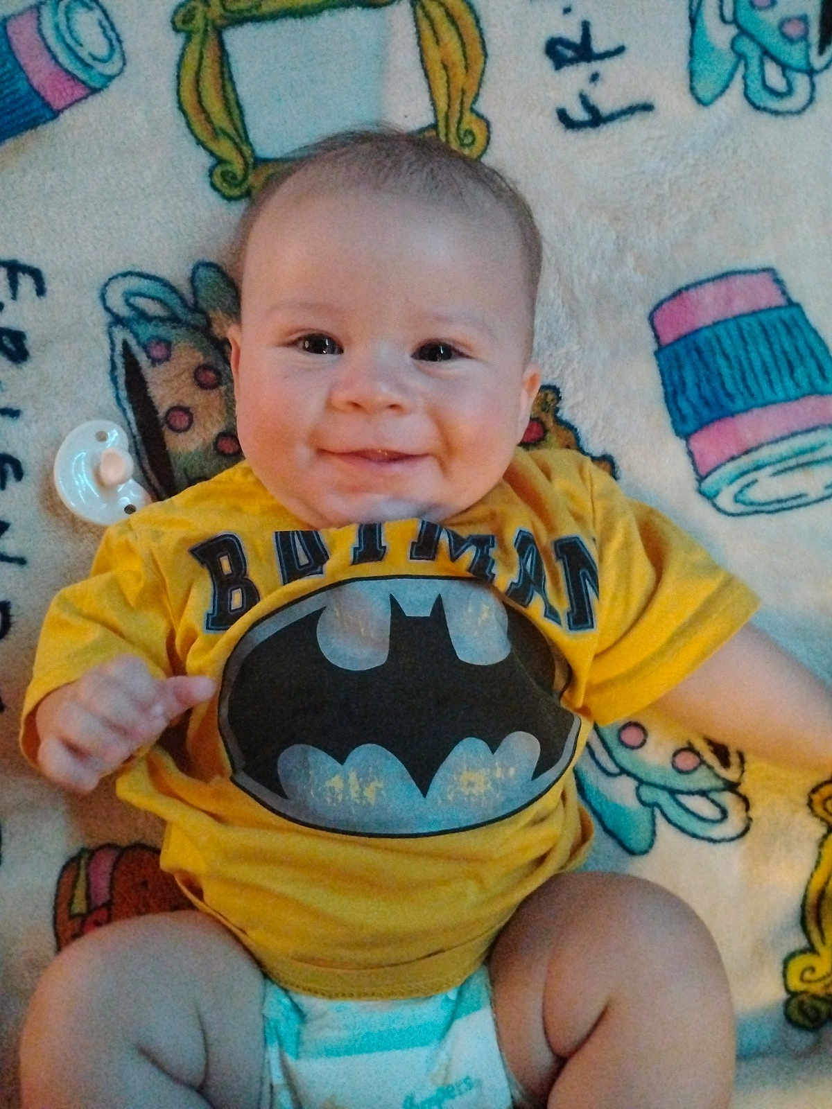 Riott is registered to the contest to win money with this photo: baby, child, smile, batman, yellow_shirt, diaper, blanket, pacifier, cute, infant, sitting, happy, chubby_cheeks, indoor, person, clothing, face, head, young, playful