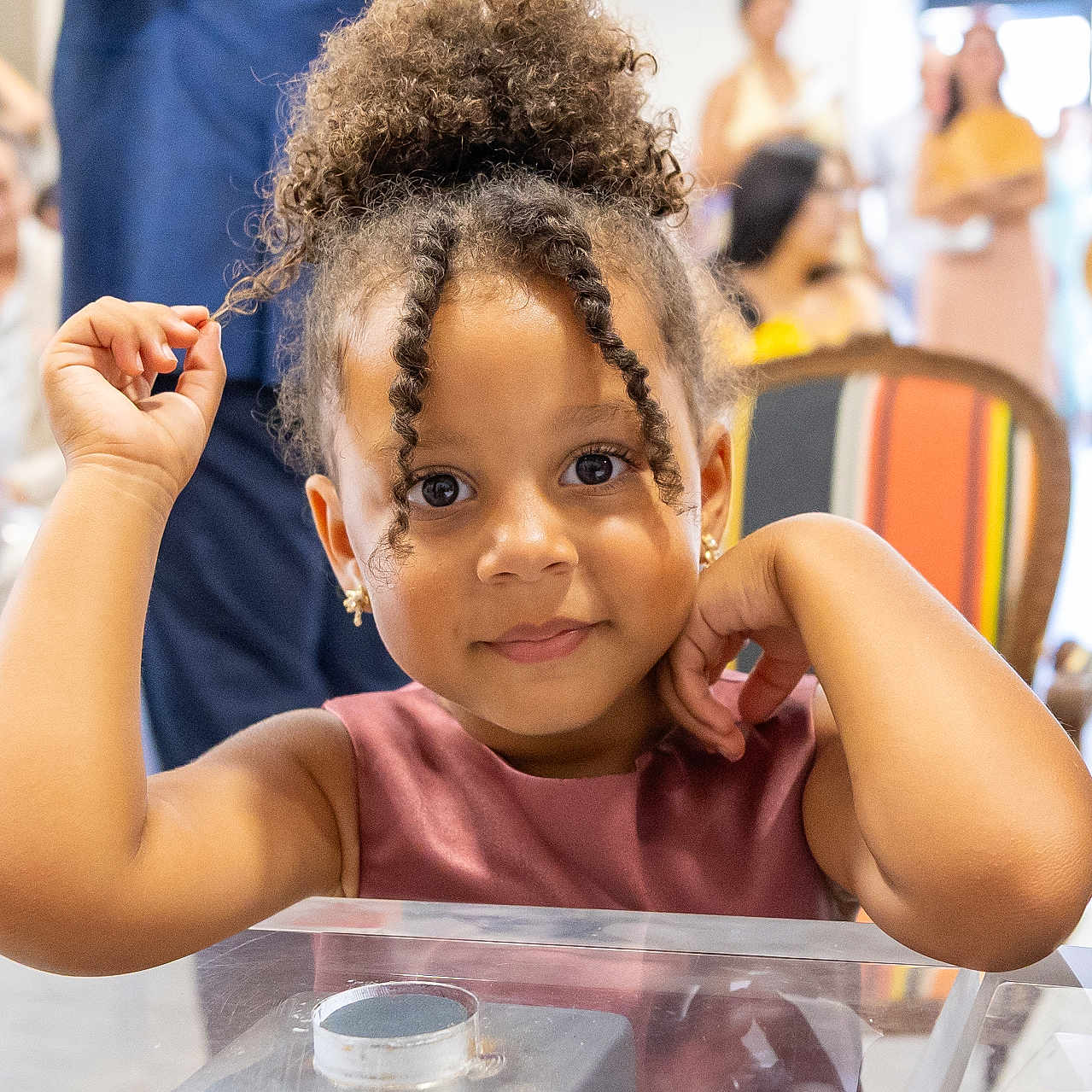 Gelsomina participe au concours pour gagner de l'argent avec cette photo : big_eyes, bun, child, colorful_chair, curly_hair, dress, earrings, event, gesture, girl, glass_surface, indoor, people_blurred, portrait, ringlet, seated, skin_tone, smile, table, toddler