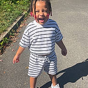 Gelsomina a rejoint le concours — aidez-le/la à gagner de superbes lots ! child, toddler, smiling, braided_hair, beaded_hairstyle, striped_shirt, striped_shorts, white_sneakers, outdoor, pavement, shadow, greenery, sunlight, happy, playful, driveway, parked_car, casual_clothing, summer, portrait