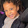 child, smiling, curly_hair, car_seat, backseat, headrest, knitted_sweater, puffer_jacket, earring, teeth, closeup, portrait, playful, happy, window, zipper, seat, cheeks, brown_eyes, candid