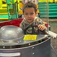 Youness joined the competition — help win amazing prizes! child, kid, steering_wheel, kiddie_ride, minion, toy, amusement, playground, indoor, colorful, silver, hoodie, curly_hair, vehicle, fun, entertainment, focus, expression, hands, face