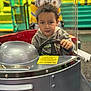 child, kid, steering_wheel, kiddie_ride, minion, toy, amusement, playground, indoor, colorful, silver, hoodie, curly_hair, vehicle, fun, entertainment, focus, expression, hands, face