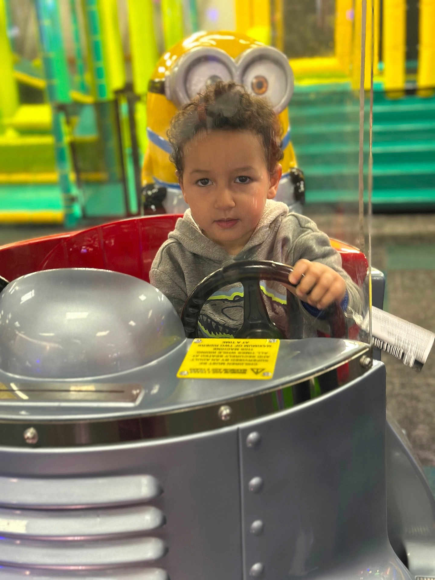 Youness joined the competition — help win amazing prizes! child, kid, steering_wheel, kiddie_ride, minion, toy, amusement, playground, indoor, colorful, silver, hoodie, curly_hair, vehicle, fun, entertainment, focus, expression, hands, face