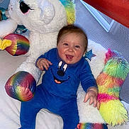 Tiago a rejoint le concours — aidez-le/la à gagner de superbes lots ! baby, blue_onesie, child, colorful, cozy, cute, face, fun, happy, indoor, infant, person, playpen, plush_toy, rainbow_colors, sitting, smiling, soft_toys, toy, unicorn