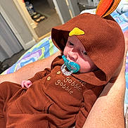 Noah joined the competition — help win amazing prizes! baby, child, pacifier, hood, turkey_costume, brown_clothing, indoor, blanket, person, arm, hand, cute, infant, sleepy, fabric, button, patterned_blanket, bedroom, comfort, resting
