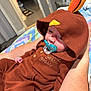 baby, child, pacifier, hood, turkey_costume, brown_clothing, indoor, blanket, person, arm, hand, cute, infant, sleepy, fabric, button, patterned_blanket, bedroom, comfort, resting