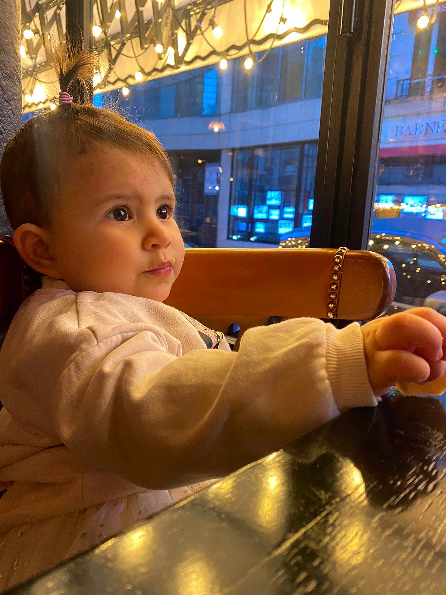 Alaïa-Victoria a rejoint le concours — aidez-le/la à gagner de superbes lots ! toddler, child, pony_tail, indoor, table, wooden_table, window, city_lights, reflection, warm_lighting, clothing, long_sleeves, face, expression, curious, seated, restaurant, evening, urban, chair