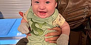 Maialen is registered to the contest to win money with this photo: baby, smiling, child, green_bow, green_outfit, mittens, adult, leather_jacket, indoor, window_blinds, happy, portrait, person, holding, seated, cute, infant, clothing, face, hands