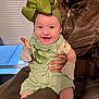baby, smiling, child, green_bow, green_outfit, mittens, adult, leather_jacket, indoor, window_blinds, happy, portrait, person, holding, seated, cute, infant, clothing, face, hands