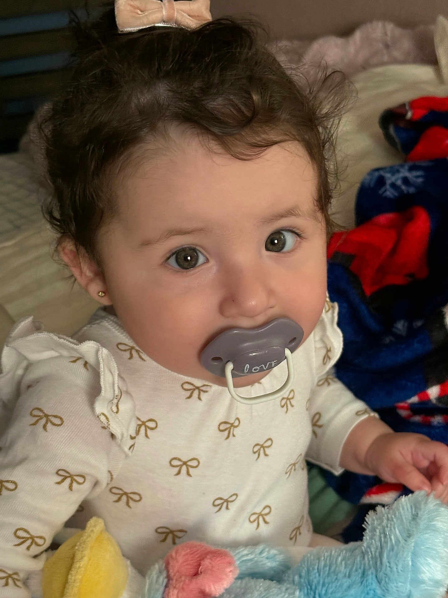 Maialen is registered to the contest to win money with this photo: baby, pacifier, bow, curly_hair, blanket, plush_toy, infant, cute, child, indoors, soft_toys, clothing, earrings, portrait, looking_at_camera, sitting, long_sleeve, face, hands, cozy
