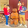 family, children, child, toddler, parents, outdoor, cabin, wooden_house, red_wall, bear_statue, wood_carving, group_portrait, smiling, standing, holding_child, window, firewood, wooden_floor, daylight, vacation