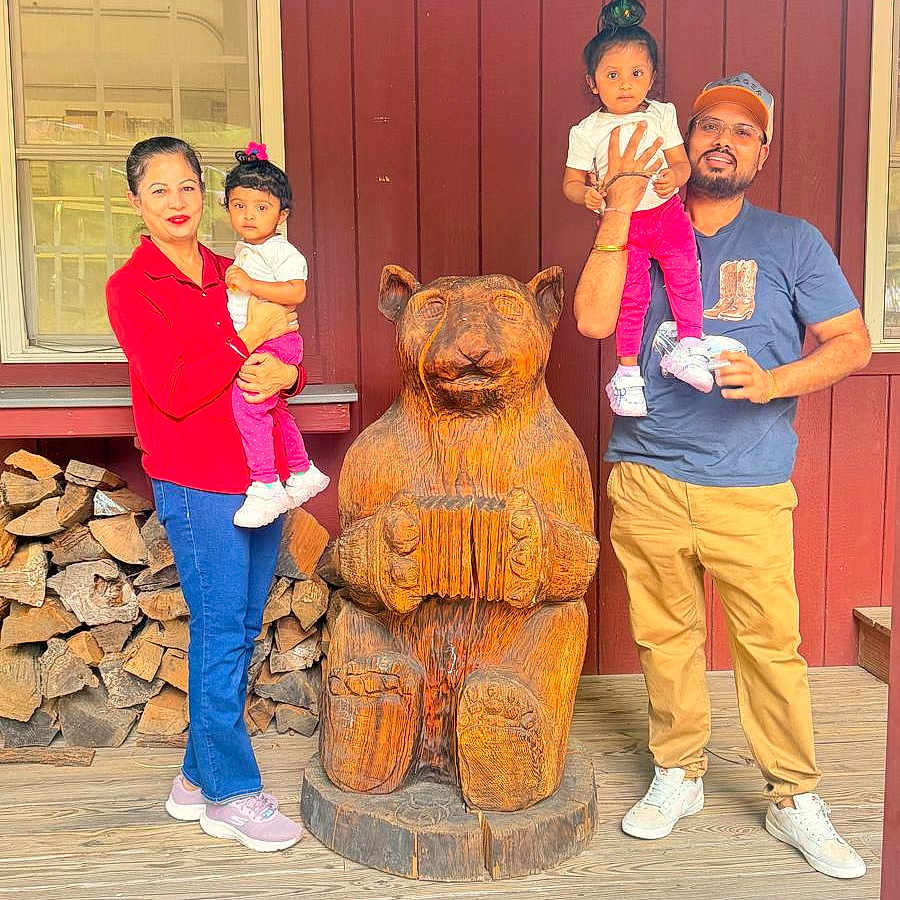 Hargun is registered to the contest to win money with this photo: family, children, child, toddler, parents, outdoor, cabin, wooden_house, red_wall, bear_statue, wood_carving, group_portrait, smiling, standing, holding_child, window, firewood, wooden_floor, daylight, vacation