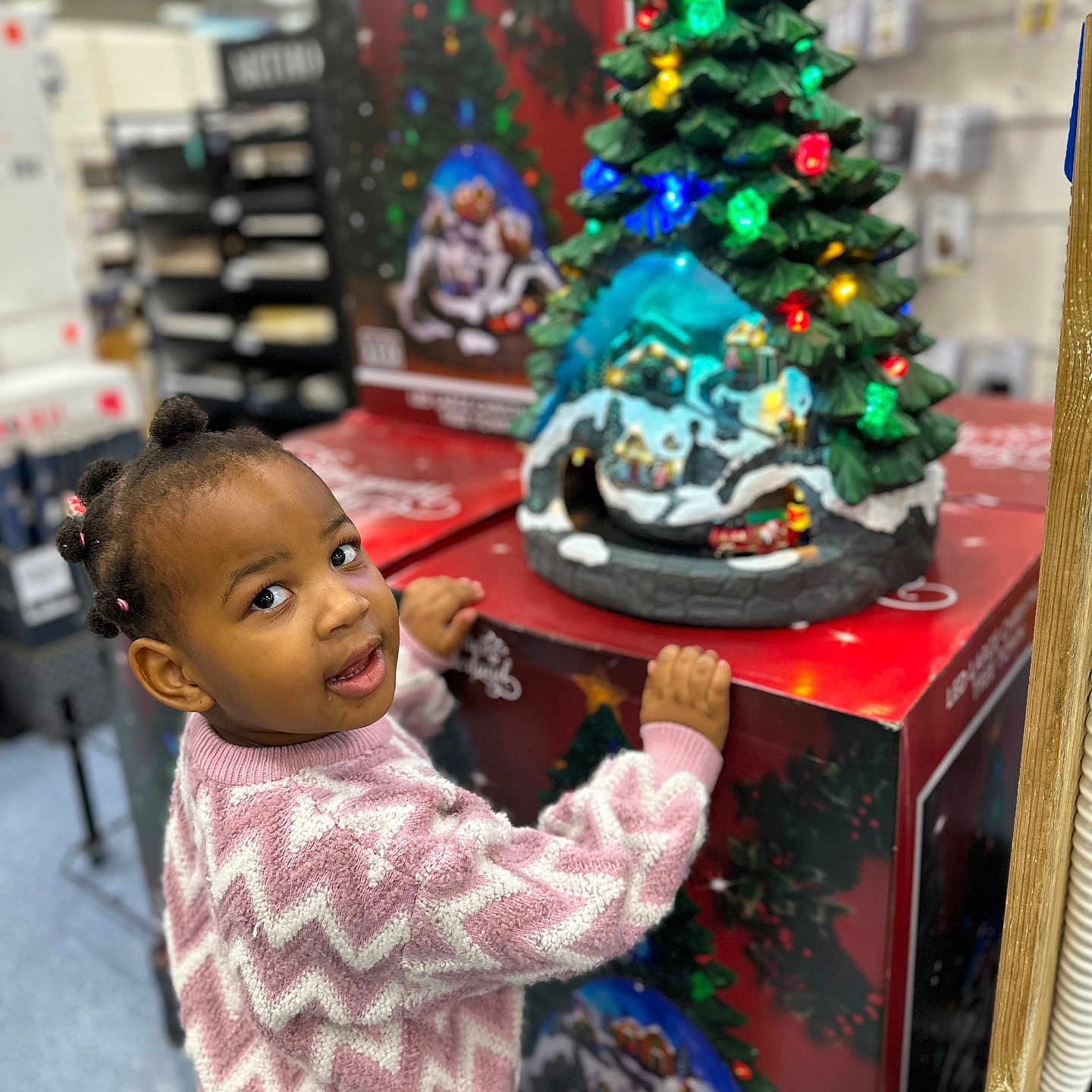 Aletheia is registered to the contest to win money with this photo: child, christmas_tree, curious, festive, girl, hair_buns, holiday_decor, indoor, lights, person, pink, retail, seasonal, shopping, smiling, store, sweater, toddler, toy, zigzag_pattern