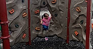 Gracielynn is registered to the contest to win money with this photo: child, playground, climbing_wall, rock_climbing, play, outdoor, trees, wood_chips, clothing, pink_shirt, blue_pants, footwear, climbing_holds, play_equipment, nature, fun, activity, daylight, person, recreation