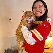 Mazie is registered to the contest to win money with this photo: woman, cat, orange_cat, red_jersey, smiling, indoor, lamp, pet_house, long_hair, holding, furry, cozy, table, happy, casual_clothing, sports_jersey, light_skin, home, expression, portrait