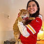 woman, cat, orange_cat, red_jersey, smiling, indoor, lamp, pet_house, long_hair, holding, furry, cozy, table, happy, casual_clothing, sports_jersey, light_skin, home, expression, portrait
