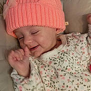 Charlie participe au concours pour gagner de l'argent avec cette photo : baby, infant, pink_hat, knit_hat, bow, heart, floral_onesie, cushion, smiling, happy, sleepy, cute, child, portrait, cozy, indoor, soft_lighting, relaxed, small_hand, lying_down