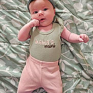 Lilith joined the competition — help win amazing prizes! baby, infant, headband, clothing, blanket, pattern, lying_down, cute, pink_pants, green_shirt, text, hand_near_mouth, face, skin, person, portrait, young_child, soft_lighting, indoor, baby_girl