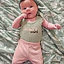 baby, infant, headband, clothing, blanket, pattern, lying_down, cute, pink_pants, green_shirt, text, hand_near_mouth, face, skin, person, portrait, young_child, soft_lighting, indoor, baby_girl