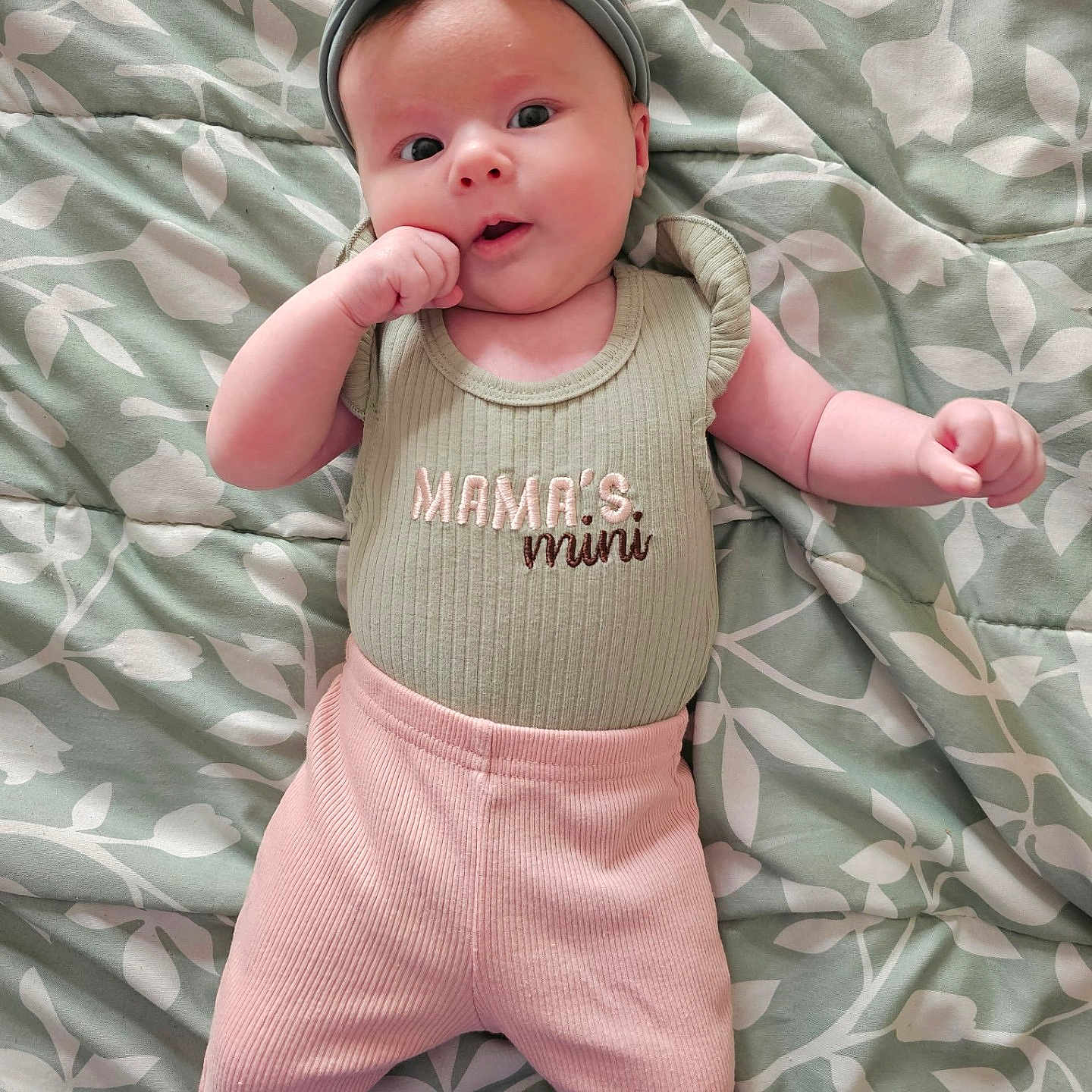 Lilith joined the competition — help win amazing prizes! baby, baby_girl, blanket, clothing, cute, face, green_shirt, hand_near_mouth, headband, indoor, infant, lying_down, pattern, person, pink_pants, portrait, skin, soft_lighting, text, young_child