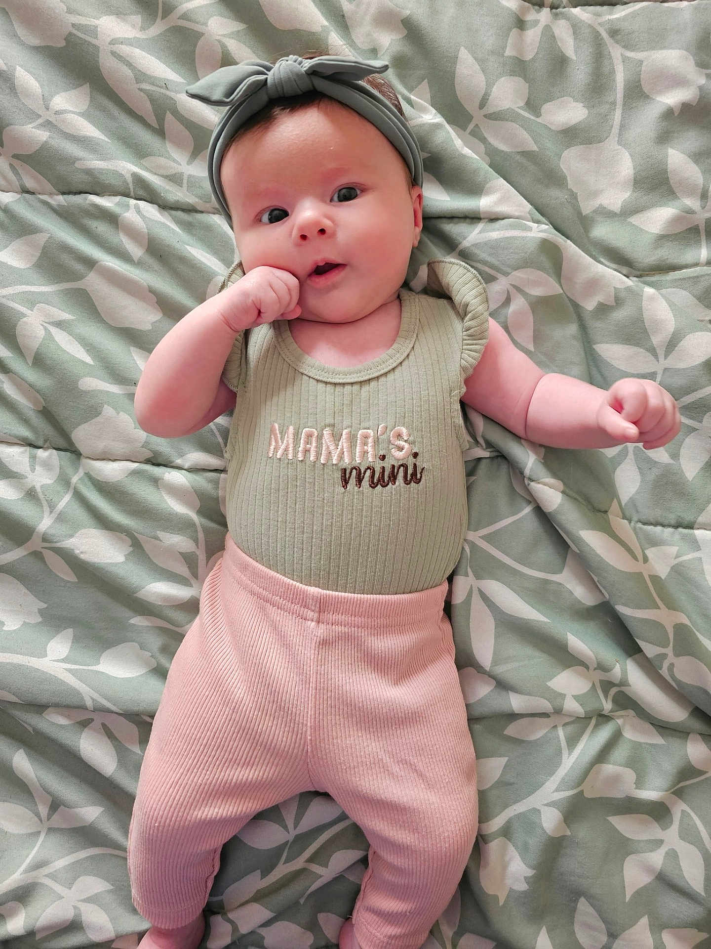 Lilith joined the competition — help win amazing prizes! baby, infant, headband, clothing, blanket, pattern, lying_down, cute, pink_pants, green_shirt, text, hand_near_mouth, face, skin, person, portrait, young_child, soft_lighting, indoor, baby_girl