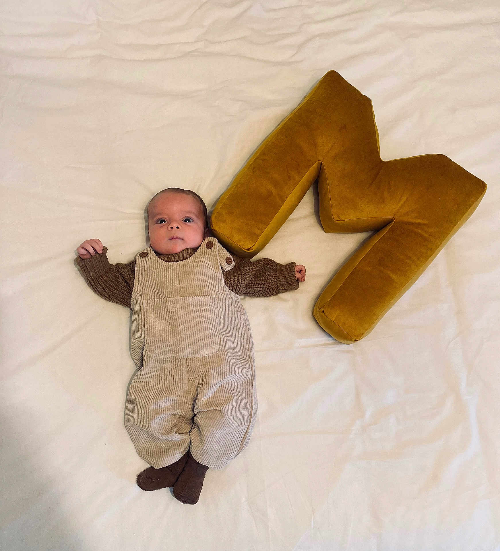 Marso participe au concours pour gagner de l'argent avec cette photo : baby, infant, plush, letter_m, blanket, brown_clothing, overalls, socks, lying_down, soft_texture, indoor, child, cute, portrait, cozy, fabric, button, head, face, small_hands
