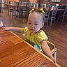 baby, child, toddler, restaurant, dining_room, table, highchair, yellow_dress, hair_bows, sitting, reaching, curious, face, portrait, wooden_floor, wooden_table, chairs, brick_wall, pendant_lights, indoor