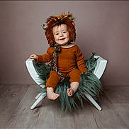 Freddie joined the competition — help win amazing prizes! baby, child, smiling, hat, costume, lion_hat, brown_outfit, chair, fluffy_cushion, green_fur, wooden_floor, indoor, portrait, cute, happy, seated, studio_photo, headwear, infant, cozy