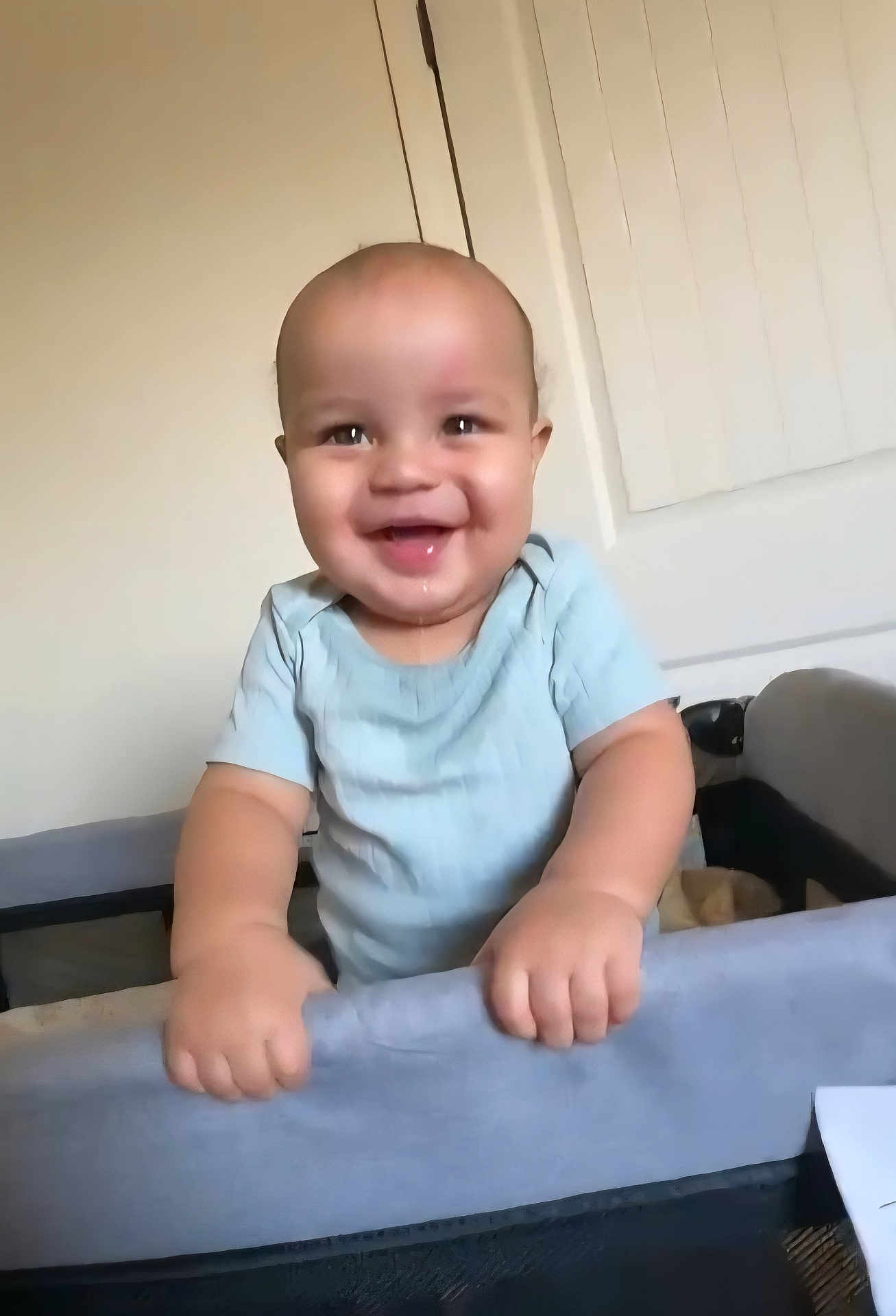 Dante is registered to the contest to win money with this photo: baby, infant, smiling, drooling, onesie, playpen, crib, hands, standing, indoor, home, cute, portrait, face, bald, chubby_cheeks, happy, door, blinds, wall