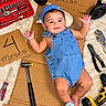 Lucas is registered to the contest to win money with this photo: baby, child, infant, smiling, portrait, lying_down, denim_overalls, blue_cap, construction_theme, tools, toolbox, cardboard, wrench, hammer, screwdriver, drill, workshop, indoor, playful, happy